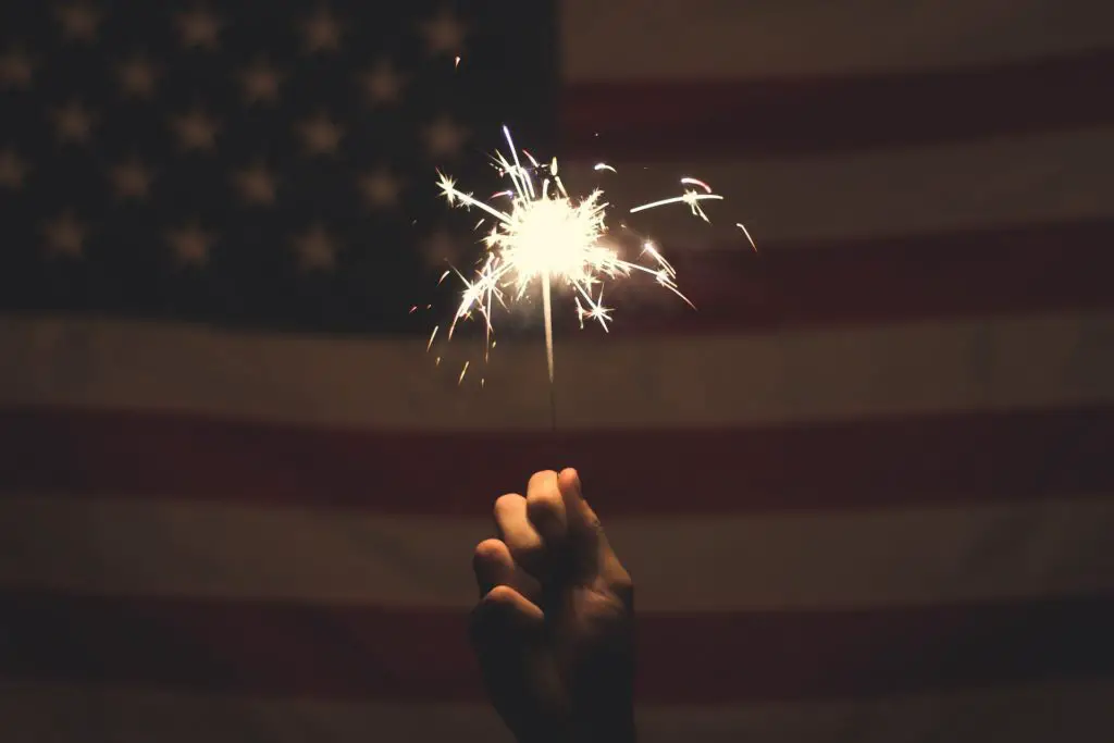 A hand holding a sparkler in front of an American flag.