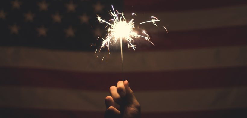 A hand holding a sparkler in front of an American flag.