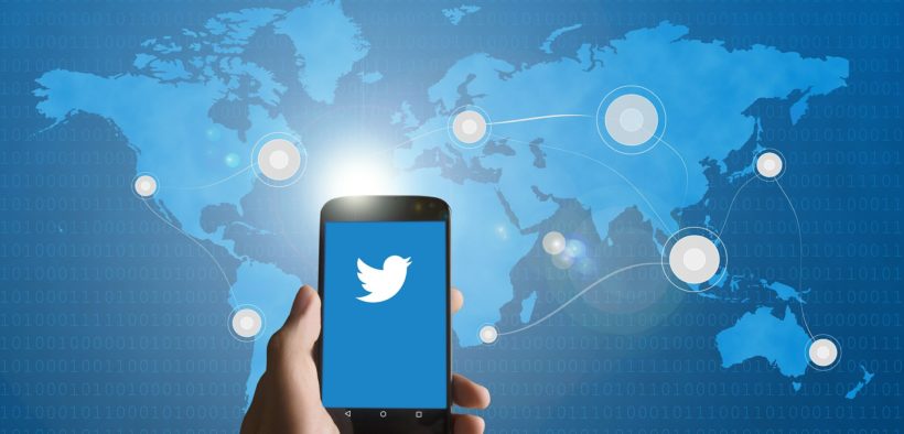Hand holding a mobile phone displaying Twitter symbol superimposed over a map of the world.
