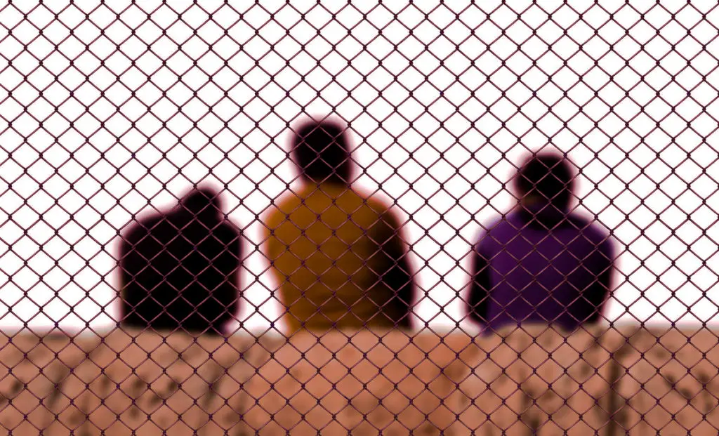 Three men sitting behind a chain link border fence.