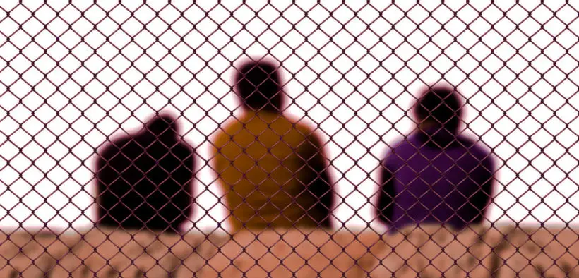 Three men sitting behind a chain link border fence.
